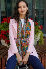 Medium full shot of brunette lady with a black handbag in a pink coat sitting on a bench in urban...