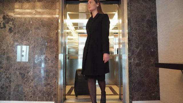 Woman carrying a suitcase comes out of the elevator and walking down the hotel lobby.