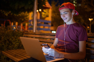 Smiling teenage boy typing message on smartphone. Handsome young man with notebook, outdoors. Cheerful guy holds a laptop on the knees and looking to the screen. Teenager in headphones with laptop