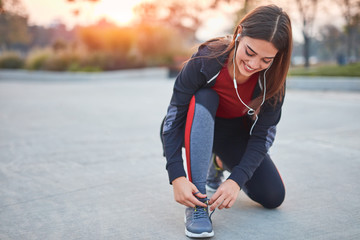 Young modern woman tying running shoes in urban park. - Powered by Adobe