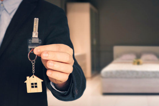 Man shows keys with keychain in the form of a little house into a new flat on blurred background of the bedroom room with a modern interior