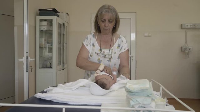 Senior midwife changing diapers newborn child on bed in hospital room at maternity ward.