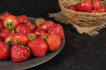 Lot of whole fresh red strawberry on gray ceramic plate in round rattan bowl with jute fabric on grey stone