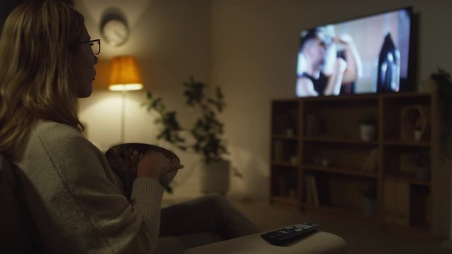 Medium shot of woman in glasses sitting on couch in living room lit by lamplight and watching fitness show on TV while eating popcorn