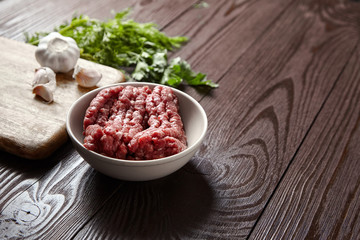 Bowl with raw minced meat, cutting board with fresh seasonings on a wooden background. White garlic heads and cloves, green dill and parsley leaves. Ground beef and condiments
