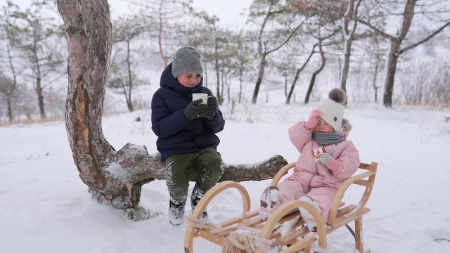 Boy drinks tea and girl eats Christmas gingerbread in jumpsuit after sledding on snowy day. Brother and sister on winter holidays together. Caring father and mother come in composition, hug, kiss kids