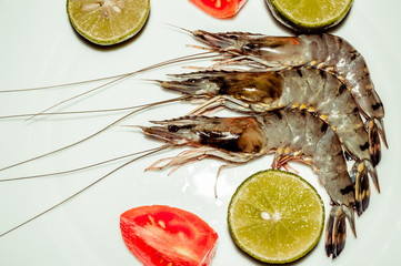 Three fresh uncooked shrimps with limes, tomatos on white plate. Raw prawns closeup. Healthy delicious seafood dinner lunch. Diet concept. Sea food background. Top view. Horizontal. Long whiskers