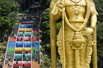 Fototapeta na wymiar (Selective focus) Stunning view of the Lord Murugan statue in the foreground and tourists climbing a colorful stairs leading to the Batu Caves in the background. Kuala Lumpur, Malaysia