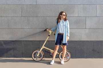 One beautiful woman stands near a stone wall and holds a folding bike, looks at the sun.