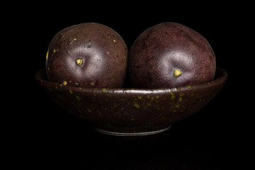 Group of two whole sweet dark purple passion fruit in dark ceramic bowl isolated on black glass