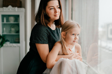 Young happy mother hugs her beautiful daughter looking at window