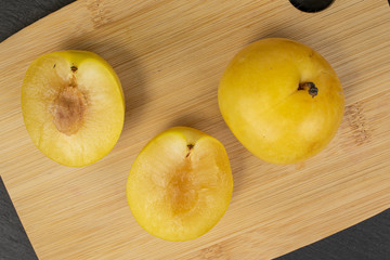 Group of one whole two halves of fresh yellow plum on bamboo cutting board flatlay on grey stone