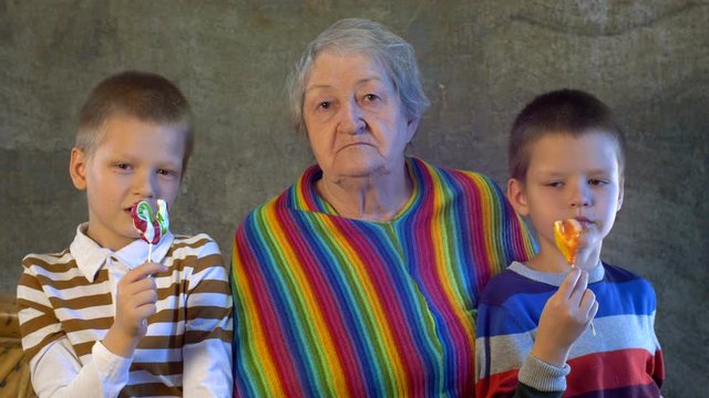 Two children in bright holiday dresses are holding caramel on stick. Children grouped around grandmother in anticipation of Christmas presents. Brothers love extravagant grandmother in bright poncho