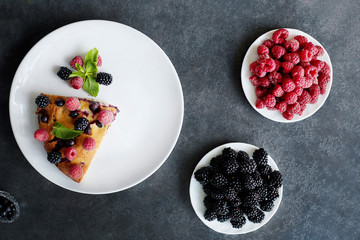 Piece of pie with blueberries, raspberry and mint for dessert on a white plate. Pieces of delicious homemade cake on a background