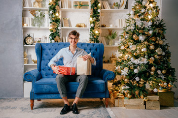 Handsome happy man with smiling face sitting on sofa with giftboxes on his legs in room with cristmas tree.