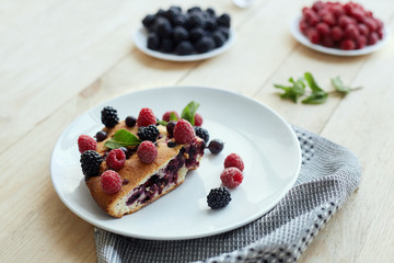 Piece of pie with blueberries, raspberry and mint for dessert on a white plate, napkin. Pieces of delicious homemade cake on a wooden boards background