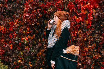 Young freckles girl in elegant clothes with cup of tea in her hand carrying funny cat inside her...