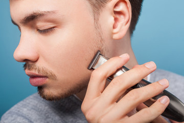 Daily shaving routine. Closeup portrait of young man grooming cheek using trimmer or electric...