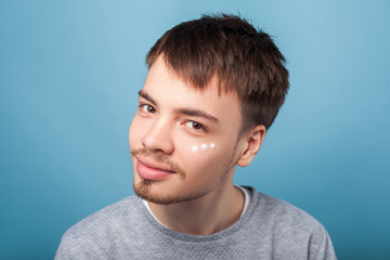 Fototapeta premium Skincare routine. Portrait of cheerful brunette man with small beard smiling at camera, having spots of moisturizer cream under eyes, male facial cosmetics. studio shot isolated on blue background