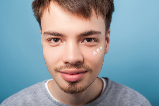 Closeup portrait of positive brunette man with small beard smiling at camera, having spots of moisturizer cream under eyes, applying facial treatment. indoor studio shot isolated on blue background