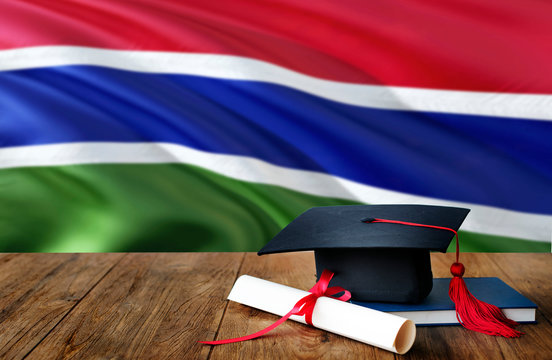 Gambia education concept. Graduation cap and diploma on wooden table, national flag background. Succesful student.