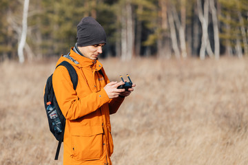 Fototapeta premium A man in an orange jacket, with a backpack and a hat, controls a quadrocopter drone outdoors in autumn.
