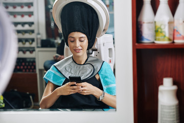 Smiling young woman reading text messages from her friends when sitting under hair steamer in beaty salon - Powered by Adobe