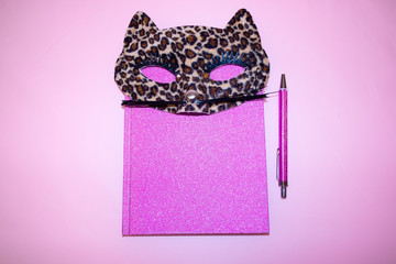 A pink diary and notebook with a cute masquerade cat mask in leopard pattern. Pink background.