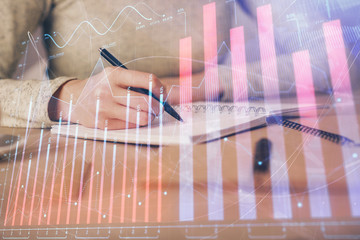 Hands writing information about stock market in notepad. Forex chart holograms in front. Concept of research. Multi exposure