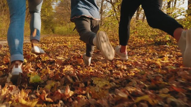 Unrecognizable two girls with a little boy running in autumn park covered with yellow leaves. Rear view close-up legs. Family enjoying the fall weather