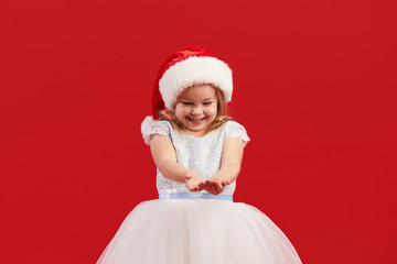 A little girl in a white princess dress and Santa hat holds snowflakes in her hands and looks at them. Red isolated background. The concept of New Year and Christmas holidays