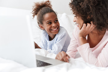 Image of african american woman and her little daughter using laptop on sofa