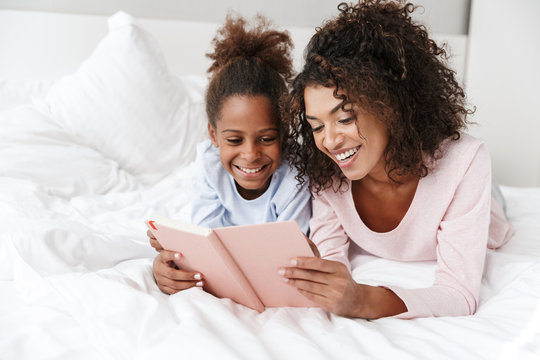 Image of african american woman and her little daughter reading book in bed