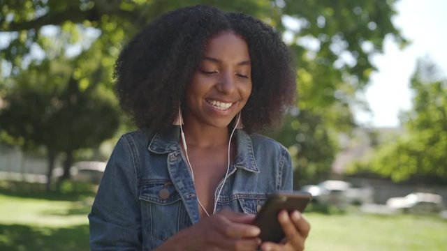 Smiling Portrait Of An African American Young Woman With Earphones In Her Ears Enjoying Chatting And Taking On Mobile Phone In Park Smiling