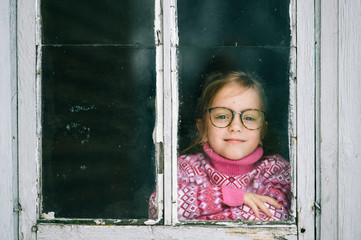 Lovely stylish little girl with happy face in glasses looking at camera through the window.
