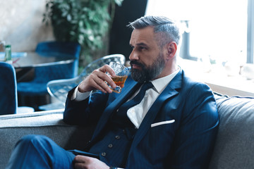 Handsome and successful businessman in stylish suit holding glass whiskey while sitting on the sofa at office.