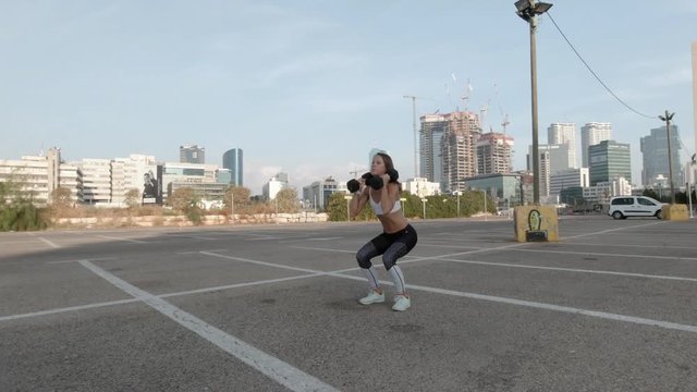 Thin muscular woman exercising with dumbbell weights as part of her fitness workout. 60fps shot in a parking lot with an urban skyline background.