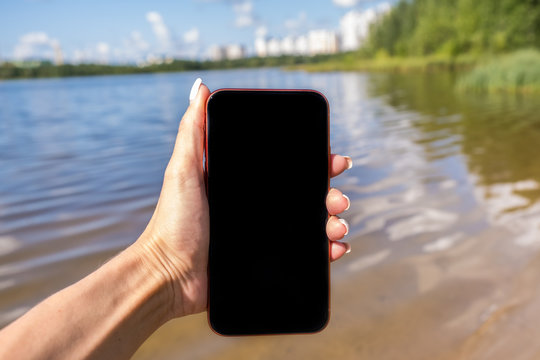 Mockup image of a woman using smart phone with blank black screen at outdoor and lake nature background.