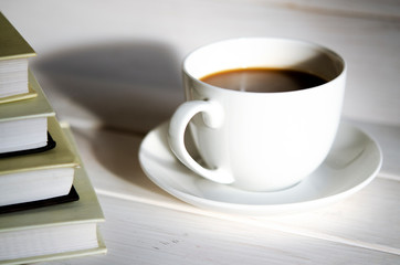 A round white cup of coffee stands on a white saucer on a white wooden table next to the shadow of the cup