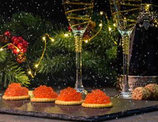 New Year and Merry Christmas concept. 2 glasses of champagne, tartlets with red caviar, spruce branches decorated with Christmas balls on a dark background.