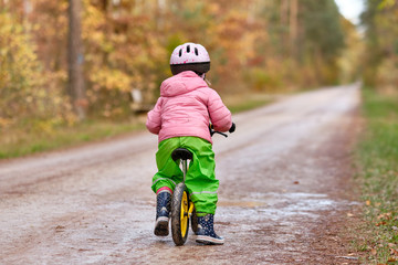 Rear view of a child girl riding on her balance bicycle in warm and waterproof clothing on a wet and dirty gravel road in the autumn forest. Seen in Franconia / Bavaria in Germany in November.