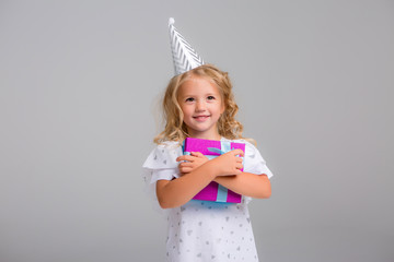little blonde girl in birthday cap smiling with gift box and confetti on white background, little beautiful girl wearing birthday cap on isolated background smiling with happy face, space for text
