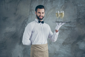 Photo of attractive handsome man with hand behind his back smiling toothily with bristle on face holding tray with glasses of champagne isolated grey concrete wall color background