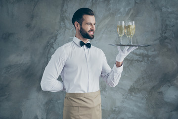 Turned photo of cheerful positive handsome waiter with hand behind his back smiling toothily holding plate with glasses of champagne isolated grey concrete wall color background