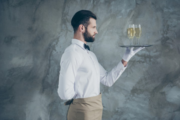 Profile side photo of attractive handsome in apron holding plate with glasses of champagne with bristle isolated grey color concrete wall background