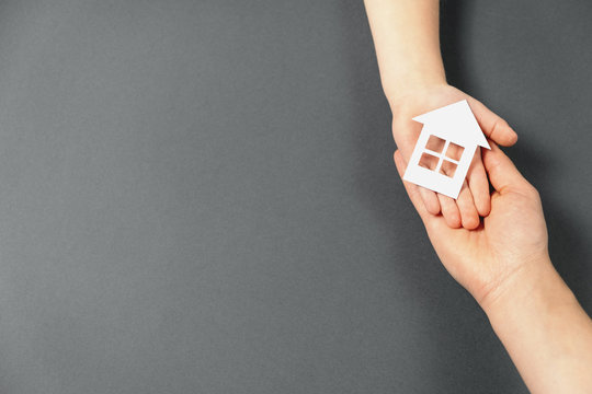 Top view on female and child hands holding white paper house on gray background. Family home and real estate concept. Flat lay, copy space