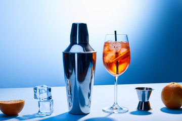 cocktail Aperol Spritz, oranges, shaker, ice cubes and measuring cup on blue background