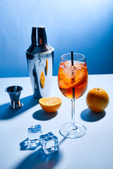 cocktail Aperol Spritz, oranges, shaker, ice cubes and measuring cup on blue background