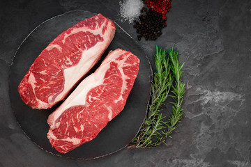 Raw striploin beef steak with rosemary, salt and pepper on round slate on stone table. Top view
