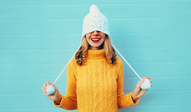 Winter portrait happy smiling young woman having fun pulls a hat over her eyes wearing yellow knitted sweater and white hat with pom pom on blue wall background